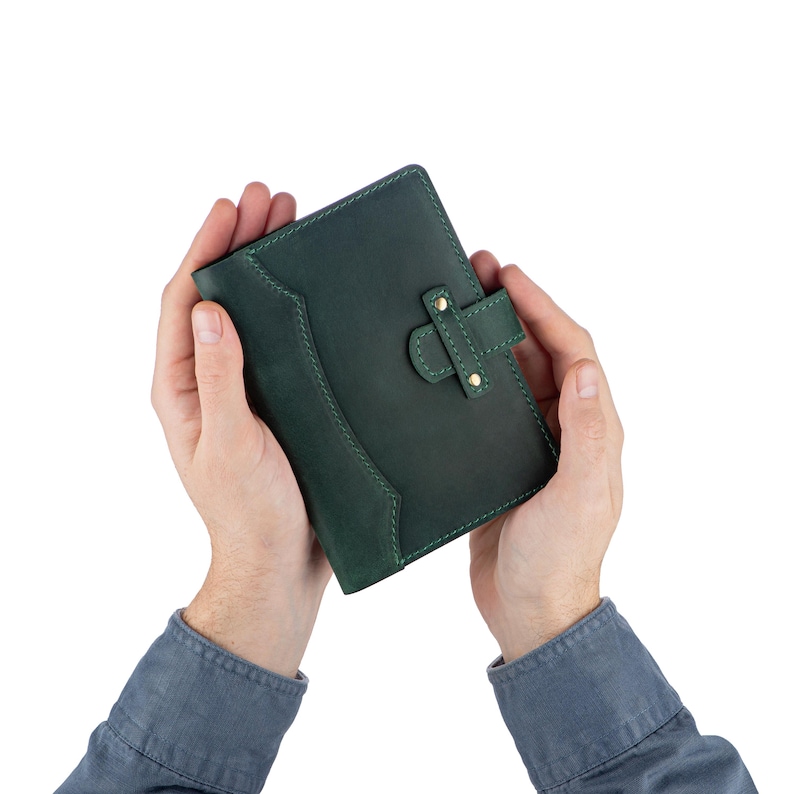 Peut inclure: Un portefeuille en cuir vert fonc&eacute; avec une fermeture &agrave; rabat et des &eacute;l&eacute;ments m&eacute;talliques de couleur laiton. Le portefeuille est maintenu ouvert, mettant en valeur son design int&eacute;rieur. Les coutures sont vert clair, et l'esth&eacute;tique g&eacute;n&eacute;rale est classique et fonctionnelle.