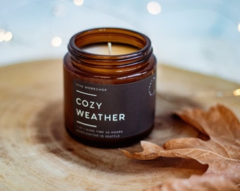 May include: A brown glass candle jar with a black label that reads "Viva Workshop Cozy Weather 20 Hour Burn Time Handcrafted in Seattle".