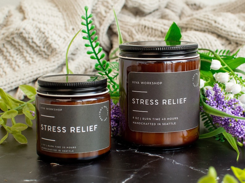May include: Two amber glass candle jars with black lids and labels. The labels read "VIVA WORKSHOP" and "STRESS RELIEF". The larger jar has a burn time of 40 hours and the smaller jar has a burn time of 30 hours. Both jars are handcrafted in Seattle.