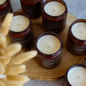 May include: A collection of amber glass candle jars with various scents, including Cranberry Prosecco, Pumpkin Spice, Morning Coffee, Fallen Leaves, Oakmoss & Amber, Leather & Smoke. The candles are arranged on a wooden surface with dried pampas grass.