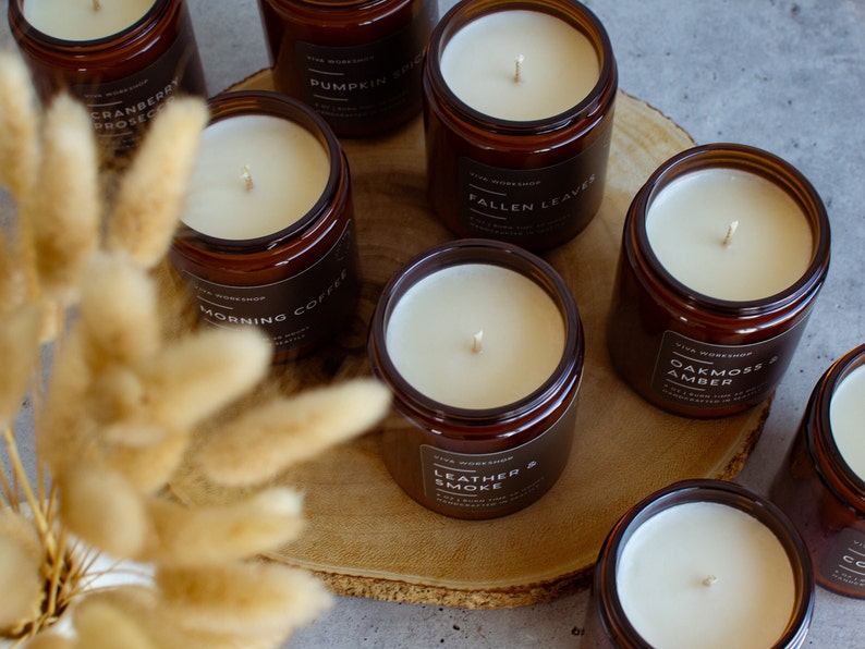 May include: A collection of amber glass candle jars with various scents, including Cranberry Prosecco, Pumpkin Spice, Morning Coffee, Fallen Leaves, Oakmoss & Amber, Leather & Smoke. The candles are arranged on a wooden surface with dried pampas grass.
