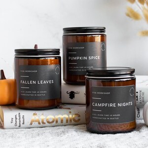 Puede incluir: Tres velas en frascos de vidrio &aacute;mbar con etiquetas negras. Las etiquetas dicen Viva Workshop, 8 oz, tiempo de combusti&oacute;n 40 horas, hecho a mano en Seattle, y los aromas Fallen Leaves, Pumpkin Spice y Campfire Nights.