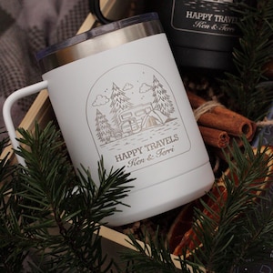 Puede incluir: Taza de viaje blanca con asa y borde plateado, con una escena de camping con &aacute;rboles, una caravana y una tienda de campa&ntilde;a. El texto "HAPPY TRAVELS Ken & Terri" est&aacute; debajo. Se ve una taza negra.