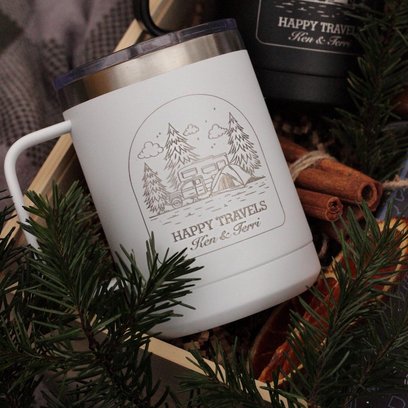 Puede incluir: Taza de viaje blanca con asa y borde plateado, con una escena de camping con &aacute;rboles, una caravana y una tienda de campa&ntilde;a. El texto "HAPPY TRAVELS Ken & Terri" est&aacute; debajo. Se ve una taza negra.