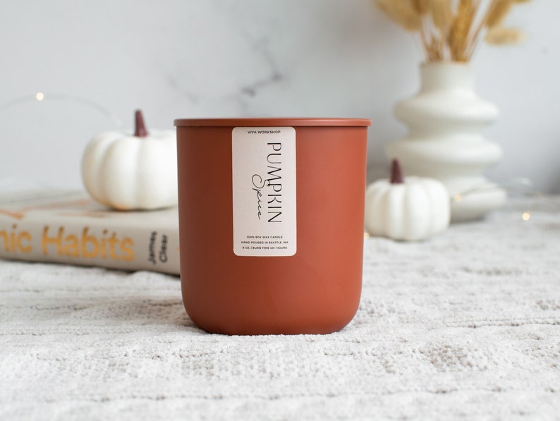 Puede incluir: Un tarro de vela de color terracota con una etiqueta blanca que dice "Viva Workshop Pumpkin Spice 100% Soy Wax Candle Hand Poured in Seattle, WA 8 oz. Burn Time 40-50 Hours"