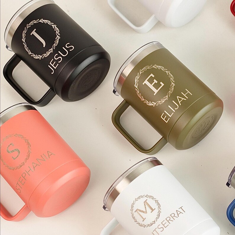 Puede incluir: Una variedad de tazas de viaje aisladas en varios colores, incluyendo negro, verde oliva, coral y blanco. Cada taza tiene un asa, un borde plateado y texto personalizado con nombres como "Jesus" y "Elijah", junto con un dise&ntilde;o de corona decorativa.
