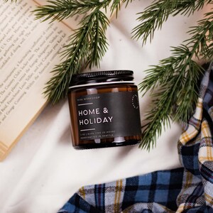 May include: A brown glass candle jar with a black label that reads "Home & Holiday" and "Vive Workshop". The candle is surrounded by a book, a plaid blanket, and pine branches.