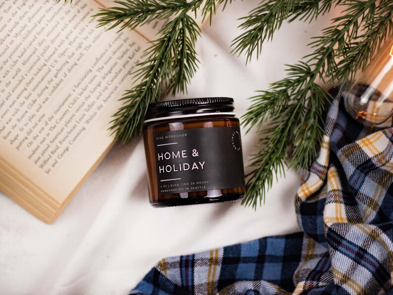 May include: A brown glass candle jar with a black label that reads "Home & Holiday" and "Vive Workshop". The candle is surrounded by a book, a plaid blanket, and pine branches.