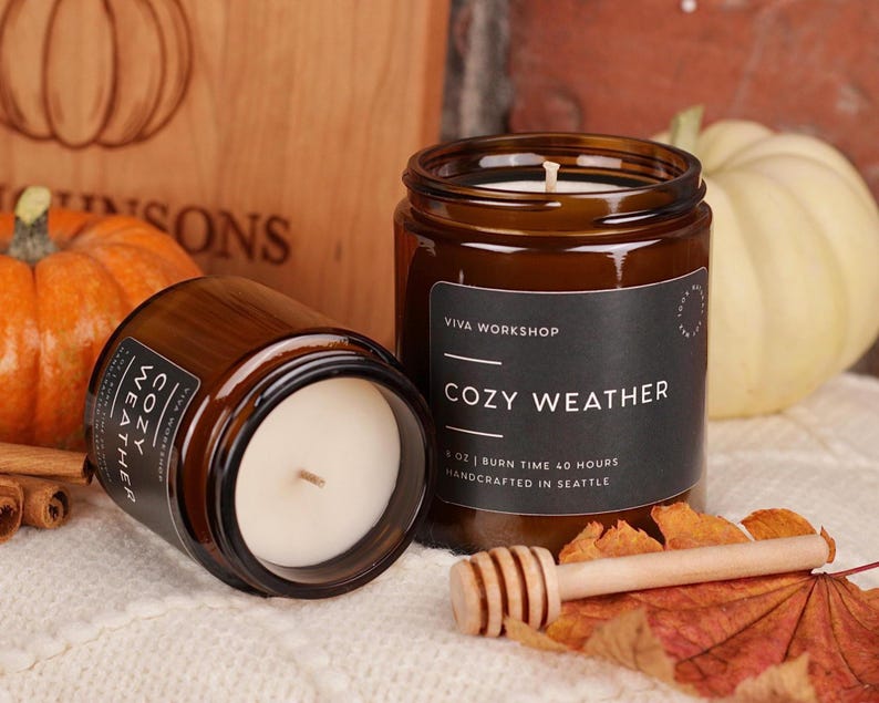 Puede incluir: Dos velas en frasco de vidrio &aacute;mbar con etiquetas negras que dicen COZY WEATHER, VIVA WORKSHOP, 8 OZ, BURN TIME 40 HOURS, HANDCRAFTED IN SEATTLE. Las velas contienen cera blanca con una mecha central, acompa&ntilde;adas de calabazas.