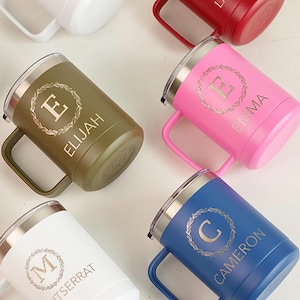 Puede incluir: Una variedad de tazas aisladas personalizadas en varios colores, incluyendo verde oliva, rosa, blanco y azul. Cada taza tiene un asa, un borde plateado y una tapa. Las tazas est&aacute;n grabadas con una letra dentro de una corona y un nombre, como "Elijah" y "Emma."
