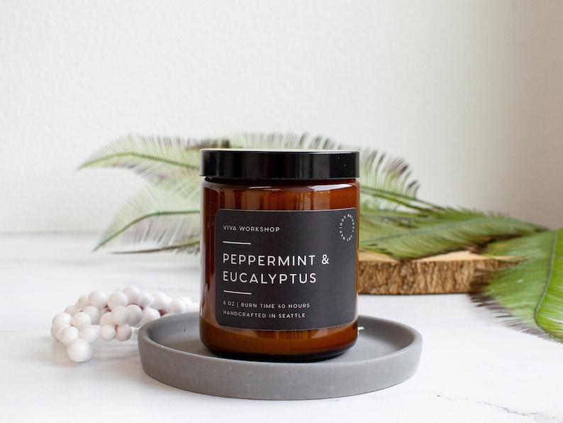 Puede incluir: Un tarro de vela de vidrio marr&oacute;n con una etiqueta negra que dice "Viva Workshop Peppermint & Eucalyptus 8 oz | Burn Time 40 Hours Handcrafted in Seattle".