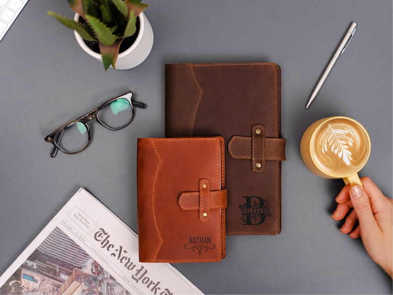 Peut inclure: Deux journaux en cuir, un marron et un beige, avec des gravures personnalis&eacute;es. Le journal marron a une fermeture &agrave; boucle. Une tasse de caf&eacute; avec du latte art, des lunettes, un stylo et un journal sont &eacute;galement sur la surface grise.