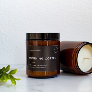 May include: Two amber glass candle jars with black lids. The jar on the left has a label that reads "VITA WORKSHOP" and "MORNING COFFEE" with the text "8 OZ | BURN TIME 40 HOURS | HANDCRAFTED IN SEATTLE". The jar on the right is filled with a white candle.