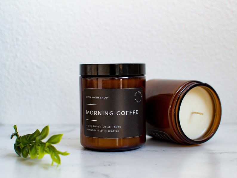 May include: Two amber glass candle jars with black lids. The jar on the left has a label that reads "VITA WORKSHOP" and "MORNING COFFEE" with the text "8 OZ | BURN TIME 40 HOURS | HANDCRAFTED IN SEATTLE". The jar on the right is filled with a white candle.