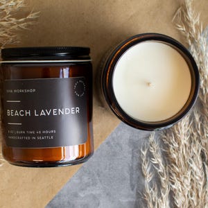 May include: Two glass jar candles with black lids, one closed and one open, sit on a neutral surface. The closed jar has a black label that reads "Beach Lavender." The open candle reveals a white wax interior. Dried plants are arranged around the candles.
