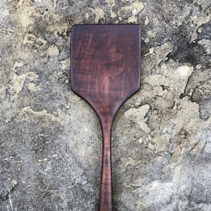 May include: A handcrafted wooden spatula with a dark brown finish. The spatula features a rectangular head and a long, slender handle. The wood grain is visible, adding to the rustic charm of the kitchen utensil.