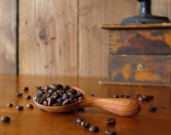 May include: A wooden spoon filled with dark brown coffee beans sits on a wooden table. Scattered coffee beans are visible around the spoon.