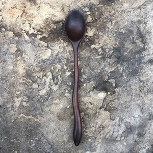 Peut inclure: Une cuill&egrave;re en bois brun fonc&eacute; avec un manche incurv&eacute;. La cuill&egrave;re est pos&eacute;e sur une surface grise textur&eacute;e.