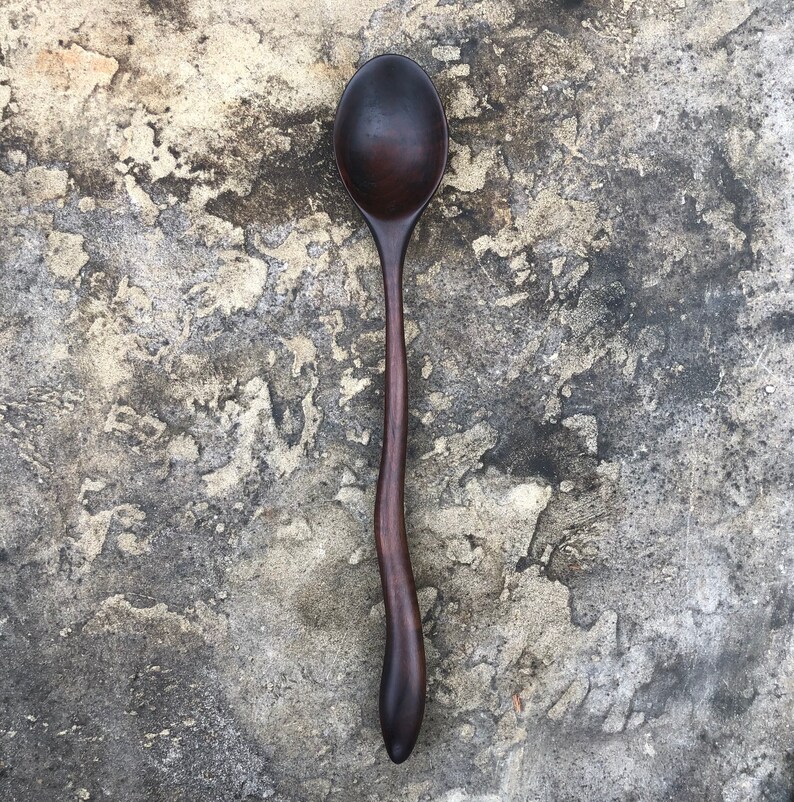 Peut inclure: Une cuill&egrave;re en bois brun fonc&eacute; avec un manche incurv&eacute;. La cuill&egrave;re est pos&eacute;e sur une surface grise textur&eacute;e.