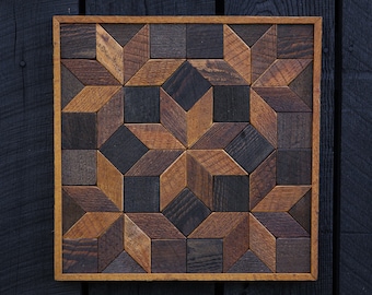 Puede incluir: Una pieza de arte de pared cuadrada de madera con un patrón geométrico de piezas de madera marrón y negro. Las piezas de madera están dispuestas en forma de estrella.