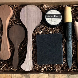 May include: A wooden box filled with brown paper shreds contains a set of wooden spoons, a black sponge, a small jar of wood oil labeled "Spoon Butter", a black handled knife with a leather sheath, and a wooden handled knife.