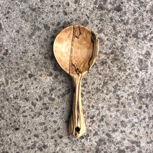Könnte beinhalten: Ein Holzlöffel mit einer natürlichen Holzoptik und einem dunkelbraunen Holzgriff. Der Löffel hat ein einzigartiges Maserungsmuster und ein kleines Loch im Griff.