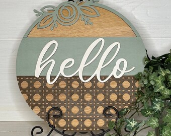 May include: Round wooden sign with the word "hello" in white script. The sign features a light brown top section with a floral design, a light green middle section, and a brown woven bottom section. It is displayed on a black metal stand.