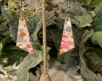 May include: A pair of abstract geometric earrings. Each earring is a light-colored, irregular triangle with a watercolor pattern of pink, orange, blue, and yellow spots. The earrings hang from silver-colored hooks.