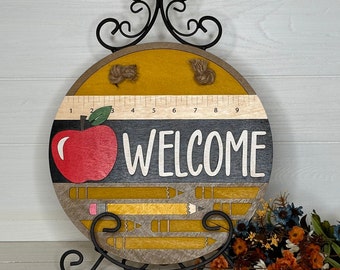 May include: Round wooden welcome sign with a black metal stand. The sign features a red apple, pencils, and the word "WELCOME" in white. A ruler is printed across the top. The sign is approximately 10 inches in diameter.