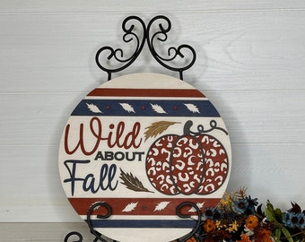 May include: A round wooden sign with the words "Wild About Fall" in burgundy script. The sign features a pumpkin with a leopard print design and is displayed on a black metal stand. Decorative fall leaves and a floral arrangement are also present.