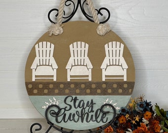 May include: Round wooden sign with three white Adirondack chairs on a tan background. The sign has a rope handle and the words "Stay Awhile" in cursive on a blue section. A decorative item for a home.