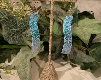 May include: A pair of abstract earrings with a wavy, elongated design. The earrings feature a teal, white, and lavender marbled pattern. They hang from silver-colored ear wires and are displayed on a burlap jewelry stand.