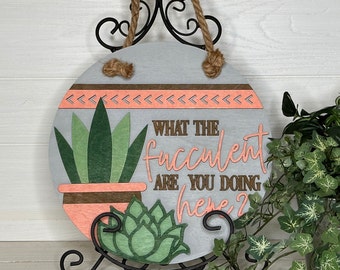 May include: A round wooden sign with a gray background, featuring a succulent plant design and the phrase "WHAT THE FUCCULENT ARE YOU DOING HERE?". The sign has a rope hanger and is displayed on a black metal stand.