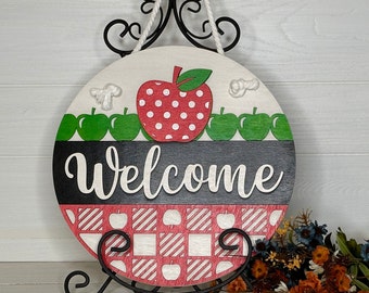 May include: A round wooden welcome sign with a red apple design and the word "Welcome". The sign features a red and white checkered pattern and a rope for hanging. It is displayed on a black metal stand.
