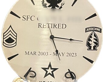 May include: A round, off-white clock with black markings and hands. The clock face features the text "SFC RETIRED" and "U.S. ARMY". It includes military insignia, a star, and the dates "MAR 2003 - MAY 2023".