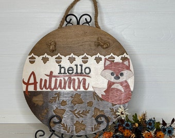 May include: Round wooden sign with a rustic design. The sign features the text "hello Autumn" and a fox illustration. The top has a jute rope for hanging. The bottom section has leaf cutouts. The sign is displayed on a black metal stand.