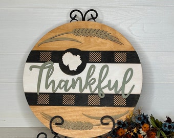 May include: Round wooden sign with a Thanksgiving theme. The sign features a natural wood grain background, a black and white buffalo plaid stripe, and the word "Thankful" in green script. A turkey silhouette is in a black circle.