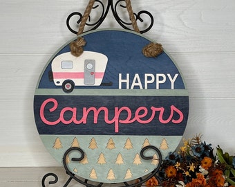May include: A round wooden sign with a blue and green background, featuring a camper illustration and the words "HAPPY Campers" in white and pink. The sign has a rope hanger and is displayed on a black metal stand. The camper has a pink door and a heart.