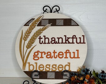 May include: A round wooden sign with the words "thankful, grateful, blessed" in brown and orange lettering. The sign has a wheat stalk design and a brown and white checkered pattern. It is displayed on a black metal stand.