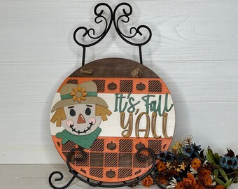May include: A round wooden fall-themed sign featuring a scarecrow design and the text "It's Fall Y'all." The sign has a brown and orange plaid pattern, a scarecrow with a green scarf, and is displayed on a black metal stand.