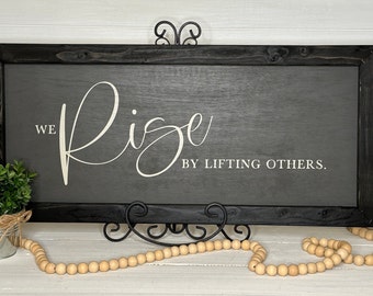 May include: A rectangular wooden sign with a black frame and a gray background. The sign features the white text "We Rise by Lifting Others." A small potted plant and a strand of wooden beads are in front of the sign.
