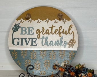May include: Round wooden sign with the words "BE grateful GIVE thanks" in a script font. The sign has a mustard yellow top, a white middle section with a decorative edge, and a teal bottom with a leaf pattern. Jute rope is attached for hanging.
