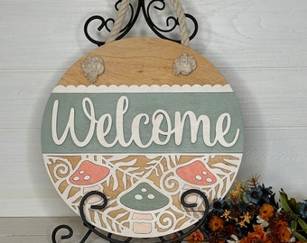 May include: A round wooden welcome sign with a rope handle. The sign features the word "Welcome" in white, with a light blue and beige background. It has a mushroom and leaf design.