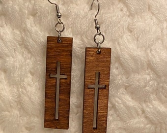 May include: A pair of wooden earrings with a rectangular shape. Each earring features a silver cross cutout in the center. The earrings hang from silver-colored fish hook ear wires.
