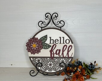 May include: A round wooden sign with the words "hello fall" in burgundy script. The sign features a floral design and is displayed on a black metal stand. The sign has a white background with a brown border.