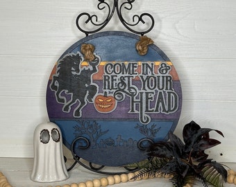 May include: A round wooden Halloween sign with the text "COME IN & REST YOUR HEAD". The sign features a headless horseman, a jack-o'-lantern, and a graveyard scene. The sign is displayed on a black metal stand with decorative accents.