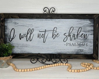 May include: A framed wooden sign with the text "I will not be shaken - PSALM 62:6" in black script. The background is white with a distressed texture. The frame is dark brown.