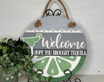 May include: A round wooden sign with a rope handle. The sign features the word "Welcome" and the phrase "HOPE YOU BROUGHT TEQUILA". A margarita glass and a lime slice are also depicted. The sign is gray, white, and green.