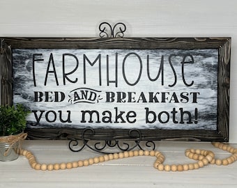 May include: A rectangular farmhouse-style wooden sign with the words "FARMHOUSE BED AND BREAKFAST you make both!" in black lettering. The sign is framed in dark brown and rests on a decorative metal stand.