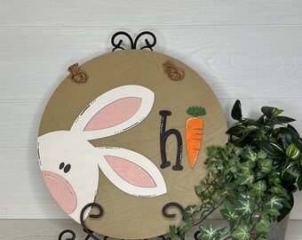 May include: A round wooden Easter sign featuring a white bunny face with pink inner ears and the word "hi" next to an orange carrot. The sign is displayed on a black metal stand and has rope for hanging.